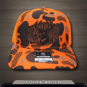 Może przedstawiać: Pomarańczowo-czarna czapka typu trucker w kamuflażu z naszywką z głową jelenia. Naszywka jest brązowa z ilustracją jelenia. Czapka ma zakrzywiony daszek i siatkowy tył. Marka "Richardson" jest widoczna na etykiecie.