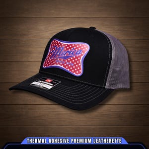 May include: Black trucker cap with a grey mesh back and a curved peak. The front displays a patch with the word "Merica" in white script, surrounded by red stars on a blue background. The text "Since 1776" is also visible.