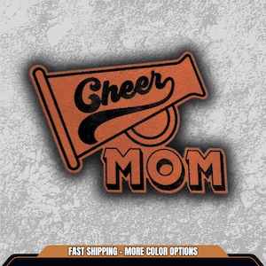 May include: An orange and black Cheer Mom design. The design features the words "Cheer" and "MOM" in a bold, stylized font, with a megaphone graphic. The design is set against a textured background.