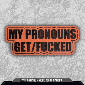 May include: An orange rectangular patch with the text "MY PRONOUNS GET/FUCKED" in black, outlined in black. The patch is set against a textured gray background.