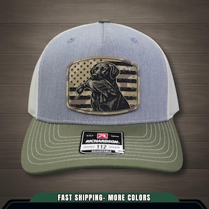 Puede incluir: Gorra de camionero gris y verde oliva con la parte trasera de malla blanca. La parte delantera presenta un parche con un perro negro sosteniendo un pato contra una bandera estadounidense de camuflaje. La gorra tiene una visera curva y una correa ajustable.