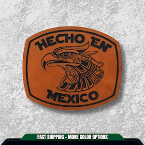 May include: A brown leather patch with a black border and the words "HECHO EN MEXICO" in black. The patch features a detailed black illustration of an eagle's head, a symbol of Mexico.