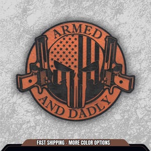 May include: A circular, orange and black patch featuring a skull design with an American flag motif. Two pistols flank the skull. The words "ARMED" and "AND DADLY" are arched around the skull. The patch is on a textured gray background.