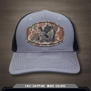 Puede incluir: Gorra de camionero gris con parte trasera de malla negra y visera curva. La parte delantera presenta un parche con un gráfico de pato en un patrón de camuflaje. La gorra tiene el texto "FAST SHIPPING- MORE COLORS" en la parte inferior.