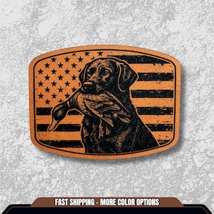 May include: A rectangular patch featuring a black Labrador Retriever holding a duck in its mouth. The background is an American flag design with stars and stripes. The patch has a distressed, vintage look.