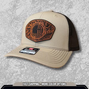 Può includere: Cappellino da camionista marrone e beige con toppa in pelle. La toppa presenta un design della bandiera americana con la scritta "THIS IS GOD'S COUNTRY". Il cappellino ha una visiera curva e un retro in rete.