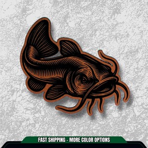 May include: A brown and black illustration of a catfish. The image features a detailed engraving style, showcasing the fish's whiskers and fins. The text at the bottom reads "FAST SHIPPING - MORE COLOR OPTIONS".