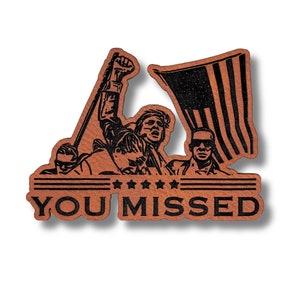 May include: Wooden pin featuring a patriotic design with an American flag and a group of people. The pin has the words "YOU MISSED" in bold letters. The pin is made of wood and has a natural wood grain finish.