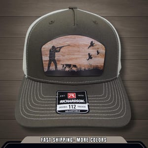 Może przedstawiać: Zielono-oliwkowa i biała czapka typu trucker z naszywką z motywem myśliwskim. Naszywka przedstawia myśliwego, psa i ptaki na tle drewna. Czapka ma zakrzywiony daszek i regulowany pasek.