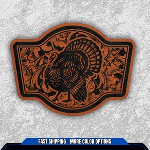 May include: A brown leather patch with a black turkey design. The turkey is surrounded by floral patterns and a black border. The patch has a unique shape and is likely for apparel or accessories.