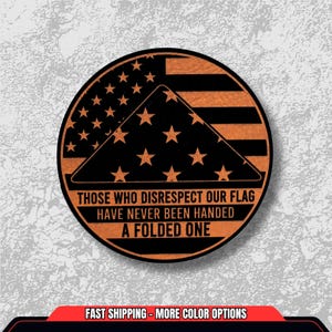 May include: A round wooden emblem featuring a stylized American flag design. The emblem includes the text: "THOSE WHO DISRESPECT OUR FLAG HAVE NEVER BEEN HANDED A FOLDED ONE." The design incorporates stars and stripes in a patriotic theme.