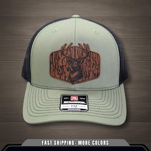 May include: A green and black trucker hat featuring a deer patch. The patch is brown with a deer head and antlers. The hat has a curved brim and a mesh back. The tag reads "RICHARDSON" and "ORIGINAL 112 TRUCKER ADJUSTABLE".
