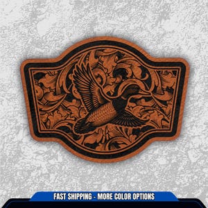 May include: A brown leather patch with a black border and a flying duck design. The duck is surrounded by floral patterns. The patch has the text "FAST SHIPPING - MORE COLOR OPTIONS" at the bottom.