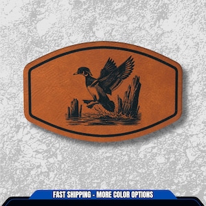 May include: A brown leather patch with a black border. The patch features a detailed illustration of a duck taking flight over water and wooden posts. The text at the bottom says "FAST SHIPPING - MORE COLOR OPTIONS".