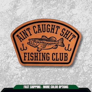 May include: A brown leather patch with the text "AINT CAUGHT SHIT FISHING CLUB" in black. A detailed illustration of a fish is in the center, with two hooks on either side. The patch has a unique shape.