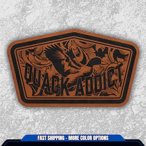 May include: A brown leather patch with a black border and the words "QUACK ADDICT" in a bold font. The patch features a detailed illustration of a duck in flight, with decorative scrollwork around the edges.