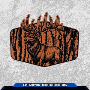 May include: An orange and black graphic of a majestic elk with large antlers, set against a textured tree bark background. The elk is facing left, with detailed linework. The image is in a hexagonal shape. The bottom of the image has the text "FAST SHIPPING - MORE COLOR OPTIONS".