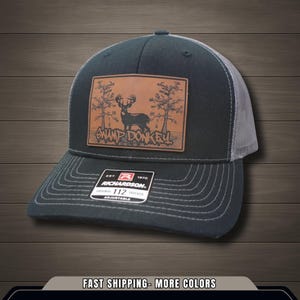 May include: A dark gray trucker hat with a light gray mesh back. The front features a brown leather patch with a deer graphic and the words "SWAMP DONKEY". The hat has a curved brim and adjustable strap.