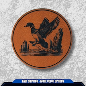May include: A round, tan leather coaster featuring a black illustration of a duck taking flight over water. The duck is depicted with its wings spread, and the image includes stylized water and wooden posts. The text "FAST SHIPPING - MORE COLOR OPTIONS" is at the bottom.