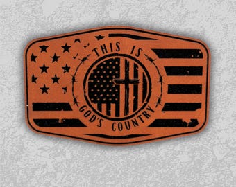 Puede incluir: Un parche rectangular de color marrón anaranjado con un diseño negro. El diseño incluye la bandera estadounidense, una cruz, alambre de púas y el texto "THIS IS GOD'S COUNTRY". Se ven estrellas y rayas.