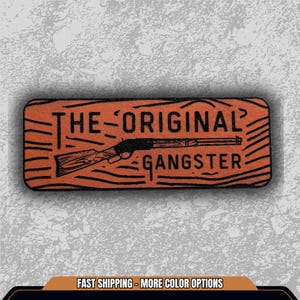 May include: Rectangular orange sticker with the text "THE ORIGINAL GANGSTER" and a black illustration of a rifle. The sticker has a wood grain texture. The text is in a bold, sans-serif font.