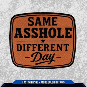 May include: A rectangular, orange-brown patch with rounded corners and black text. The text reads "SAME ASSHOLE DIFFERENT DAY" with a star in the middle. The patch has a textured appearance.