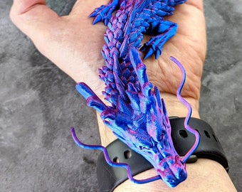May include: A blue and pink articulated dragon figurine. The dragon is held in a hand, showcasing its detailed scales and flowing form. Text on the image reads "More colours in the description" and "Blue / Pink".