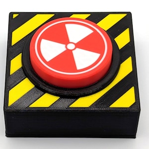 Puede incluir: Un botón cuadrado negro con rayas diagonales amarillas y negras y un botón rojo con un símbolo de radiación blanco. El botón es un artículo de novedad.