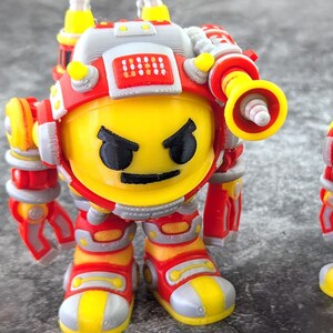 Könnte beinhalten: Zwei gelb-rote Roboterfiguren mit wütenden Gesichtern. Die Roboter haben Helme, schultermontierte Geräte und bewegliche Arme und Beine. Der größere Roboter steht vor dem kleineren. Die Roboter stehen auf einer grauen Oberfläche.