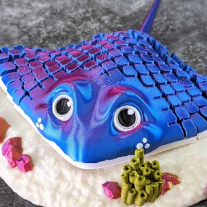 May include: A 3D printed blue and purple stingray with a white base and green coral. The stingray has a textured surface and large eyes.