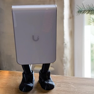 May include: A silver-colored rectangular device with a curved top and a small, stylized "U" logo, mounted on black, boot-shaped legs. The device is displayed on a wooden shelf, part of a black metal frame, with a small white vase holding greenery.