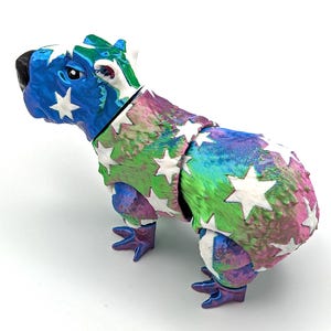 May include: A blue, green and pink capybara figurine with white stars. The figurine is made of a textured material and has a glossy finish.