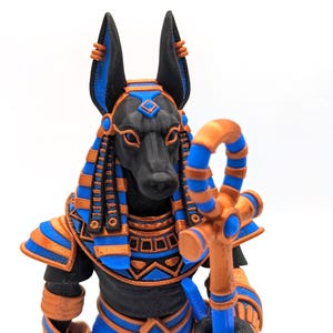 May include: A detailed figurine of Anubis, the Egyptian god of the afterlife, with a black canine head and blue and gold accents. The figure holds a staff and wears a traditional headdress. The statue is a detailed representation of ancient Egyptian art.