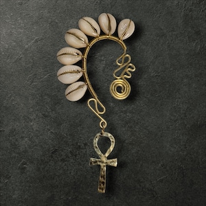 May include: A gold-toned ear cuff featuring a row of cream-colored seashells, wire detailing, and a dangling ankh charm. The ear cuff has a curved design with decorative swirls and a textured ankh pendant.