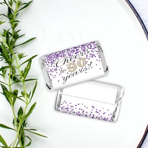 May include: Two small white chocolate bars with purple confetti and gold lettering. The text on the chocolate bar reads "Cheers to 90 years!"