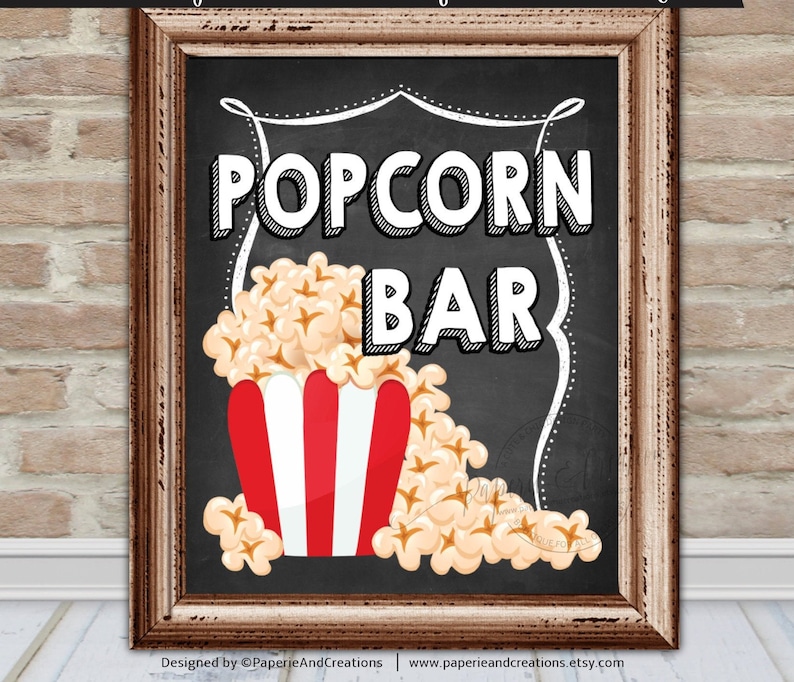 K&ouml;nnte beinhalten: Ein Schild aus Schiefer mit wei&szlig;em Rahmen, auf dem in wei&szlig;er Schrift "Popcorn Bar" steht. Ein roter und wei&szlig; gestreifter Popcornbeh&auml;lter ist mit Popcorn gef&uuml;llt und l&auml;uft &uuml;ber.