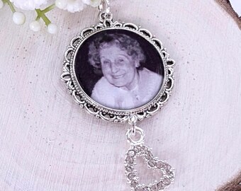 May include: A silver-toned memorial pendant with a black and white photo of a person inside an oval frame. A small, open heart charm with clear stones hangs below the frame. White flowers and a ribbon are visible.