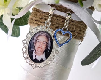 May include: A silver charm with a photo of a woman in an oval frame, and a blue heart charm, attached to a bouquet. The bouquet is wrapped in white ribbon and twine, with white flowers and greenery.
