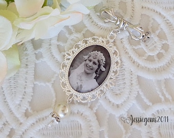 May include: A silver-toned oval pendant with a vintage black and white photograph of a woman. The pendant has a decorative filigree frame, a pearl bead, and a decorative clasp. The background is white lace.