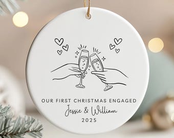May include: White ceramic Christmas ornament with a gold string. The ornament features a black line drawing of two hands toasting champagne flutes, hearts, and the text "OUR FIRST CHRISTMAS ENGAGED" with names and the year 2025.