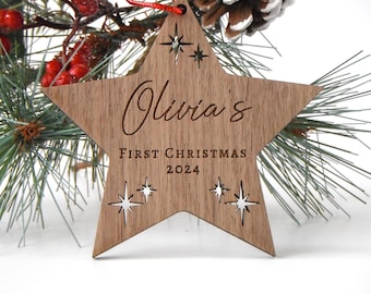 May include: A wooden star-shaped Christmas ornament with the name "Olivia's" engraved on it. The ornament also includes the text "FIRST CHRISTMAS 2024" and star-shaped cutouts. It hangs from a red string and is surrounded by pine needles and berries.