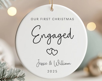 May include: White ceramic Christmas ornament with gold string. The ornament reads "OUR FIRST CHRISTMAS Engaged" in black script, with two hearts and the names "Jessie & William" and the year "2025".
