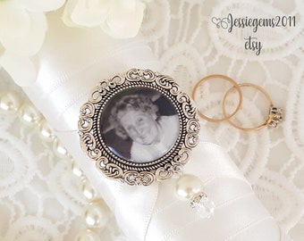 May include: A white satin ring pillow with a pearl trim and a silver locket with a black and white photo of a woman. The locket is attached to a pearl and crystal charm. Two gold wedding bands are on the pillow.