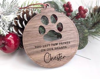 May include: A wooden Christmas ornament with a paw print cutout and the inscription "YOU LEFT PAW PRINTS ON OUR HEARTS" and the name "Chester". The ornament is brown and has a red string for hanging. The background includes pine needles, berries, and a pine cone.