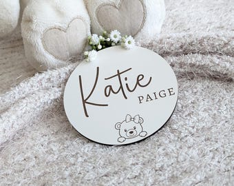 May include: A white, round wooden sign with the name "Katie" in a brown script font and the name "Paige" in a smaller, block font. A small bear illustration is at the bottom. The sign is decorated with small white flowers.