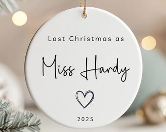 May include: White ceramic Christmas ornament with gold string. The ornament has the text "Last Christmas as Miss Hardy" in black script, a blue heart, and the year "2025". A festive holiday decoration.