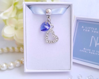 May include: A small white box with a blue ribbon and a silver charm with a blue crystal heart and a larger heart charm with clear crystals. The box has a blue label with the text "A little something for your wedding" and "Nata" in cursive. The label also says "Blue is the color of love, purity, and..."