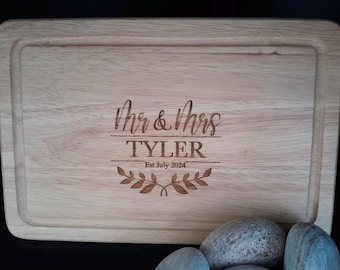 May include: A wooden cutting board with the text "Mr. & Mrs. Tyler Est. July 2024" engraved on it. The board is surrounded by a decorative border and features a leafy design.