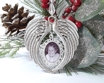 May include: Silver-toned angel wing ornament with a framed black and white photo. The ornament is suspended from a clear beaded strand. The background includes pine needles, red berries, and a pine cone, suggesting a holiday theme.