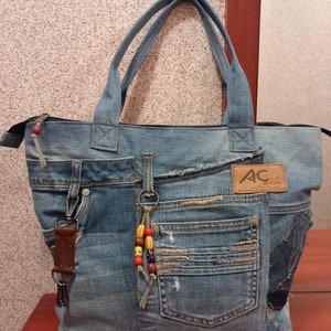 Peut inclure: Un sac cabas en jean bleu avec plusieurs poches et une fermeture éclair. Le sac a une bandoulière en cuir marron et un porte-clés avec des perles colorées. Le sac a un patch avec les lettres "AC" dessus.